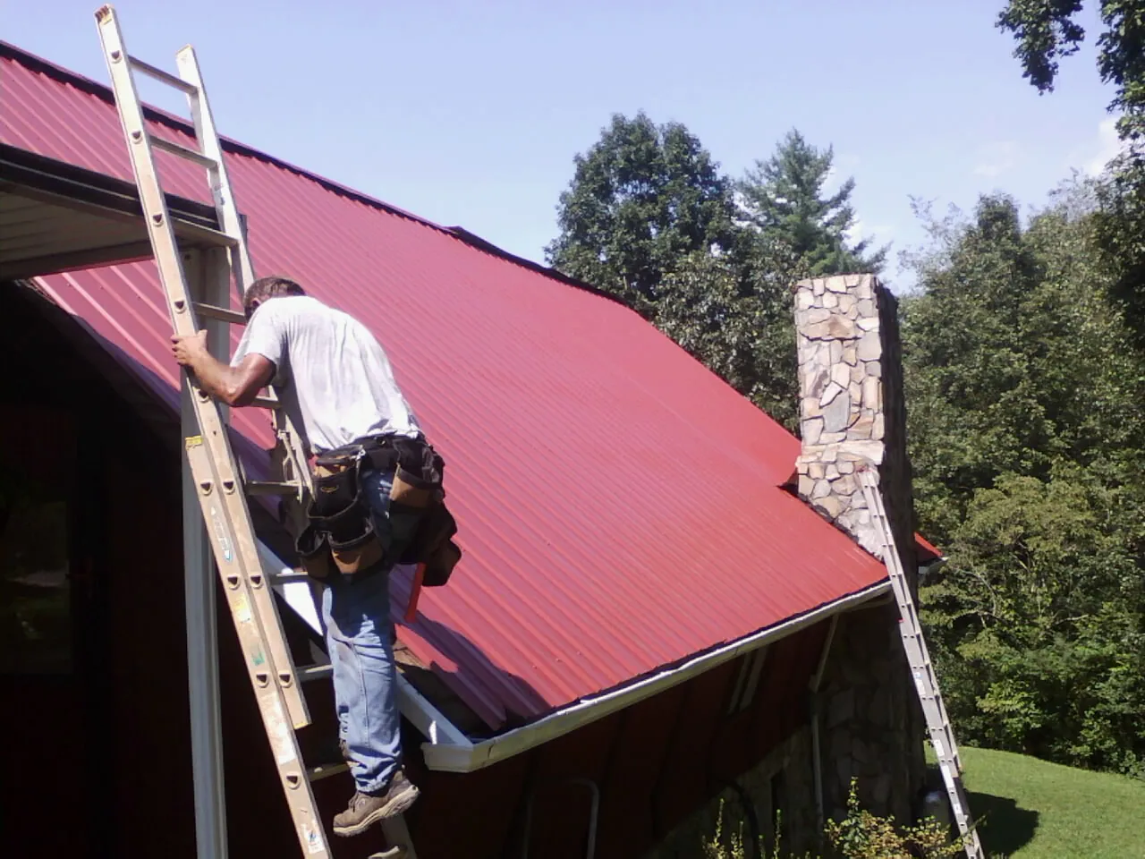 Roofer on a ladder beside a red metal roof and stone chimney — Soffit & Fascia Repair by Roofing Brothers in Irving